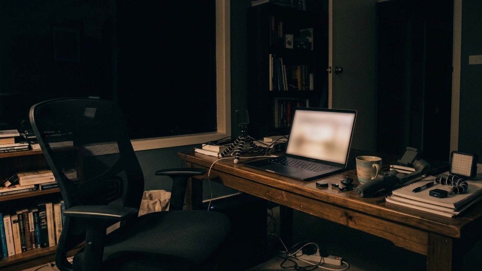 A home office in the dark, the laptop open and glowing on the desk, the chair empty, nobody in the room